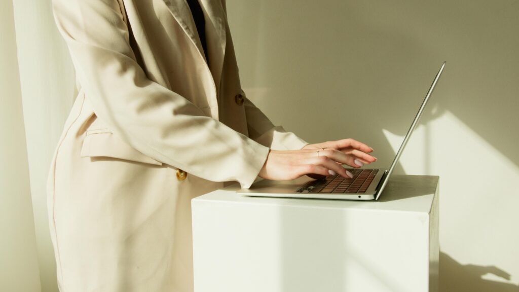 Woman in beige coat typing on laptop in a sunlit workspace, emphasizing modern work.